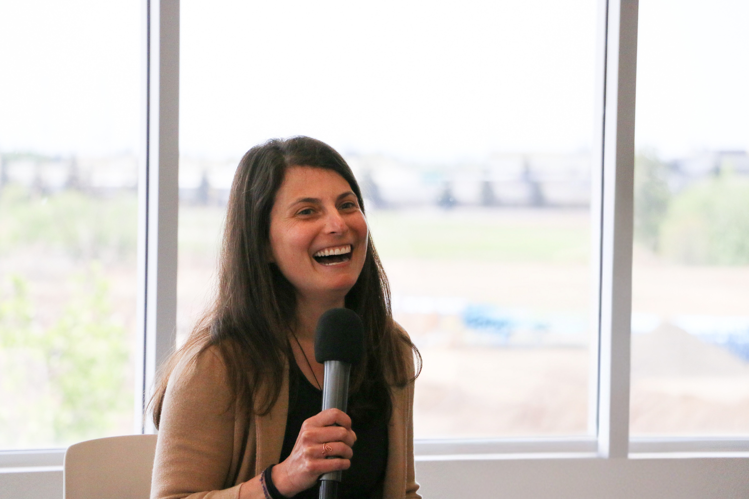 Transformational Speaker Bara Mann is on a chair in front of a window holding a microphone. She has a big smile and appears to be laughing